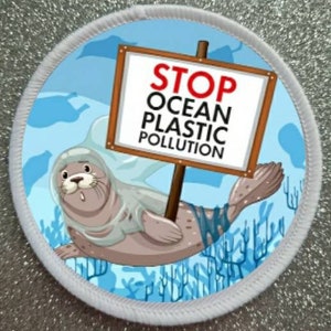 Op de afbeelding: Een ronde witte patch met een blauwe achtergrond waarop een cartoon-rob te zien is die verstrikt is geraakt in plastic zakken. De rob houdt een bord vast met de tekst "STOP OCEAN PLASTIC POLLUTION".