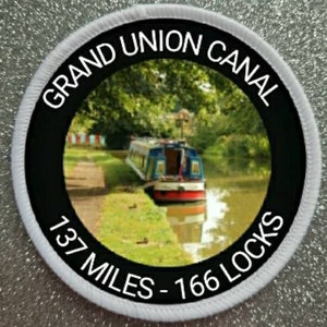 May include: A white round patch with black text and a photo of a canal with a boat. The text reads "Grand Union Canal 137 Miles - 166 Locks".