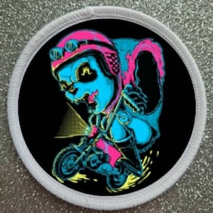 Può includere: Una toppa rotonda bianca con sfondo nero che presenta un'illustrazione al neon blu e rosa di una persona con casco e occhiali che guida una motocicletta. La motocicletta ha un faro e emette scintille.