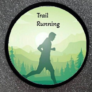 Puede incluir: Un parche circular verde y amarillo con un borde negro. El parche presenta una silueta de una persona corriendo por un bosque con el texto "Trail Running" en la parte superior.