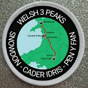 Może przedstawiać: Biała okrągła naszywka z czarną obwódką i mapą Walii. Mapa pokazuje trasę Welsh Three Peaks Challenge, która obejmuje Snowdon, Cadair Idris i Pen y Fan. Tekst "WELSH 3 PEAKS" jest napisany w czarnym kółku wokół mapy. Tekst "SNOWDON - CADER IDRIS - PEN Y FAN" jest napisany w czarnym kółku wokół mapy.