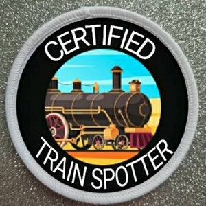Peut inclure: Un écusson rond blanc avec du texte noir qui dit "CERTIFIED TRAIN SPOTTER". Le centre de l'écusson présente une image de dessin animé d'un train à vapeur noir sur fond de ciel bleu.