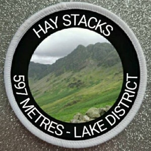 May include: A white and black round patch with the text "Hay Stacks 597 Metres - Lake District" and a photo of a green mountain range.