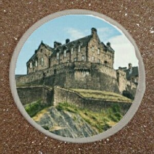 May include: A circular image of a stone castle on a hill. The castle is gray and has many windows. The hill is green and covered in grass. The image is surrounded by a white circle.