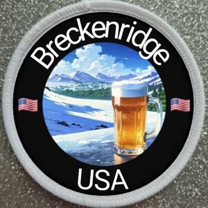 Könnte beinhalten: Ein runder weißer und schwarzer Patch mit den Worten "Breckenridge USA" und einem Bild eines Glases Bier vor einer schneebedeckten Berglandschaft mit blauem Himmel und Wolken. Auf beiden Seiten des Bildes befinden sich zwei kleine amerikanische Flaggen.