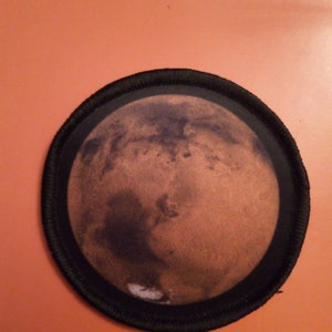 Könnte beinhalten: Ein runder, schwarz umrandeter Aufnäher mit einem detaillierten Bild des Mars, des roten Planeten, in der Mitte.