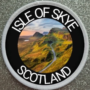 Puede incluir: Un parche circular blanco y negro con el texto "Isle of Skye Scotland" alrededor del borde. El centro del parche presenta una imagen escénica de un camino sinuoso a través de colinas verdes onduladas con una cordillera en el fondo.