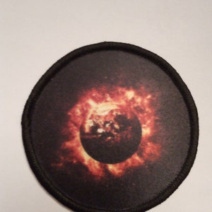 May include: A black and orange embroidered patch with a circular image of a planet with a fiery ring around it.