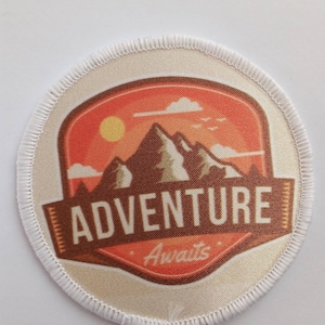 May include: A round, white, fabric patch with a brown stitched border. The patch features a colorful illustration of a mountain range with a sun and clouds. The text "ADVENTURE Awaits" is printed in brown on the patch.
