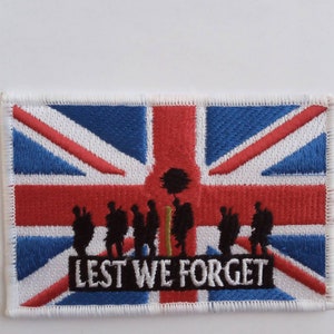 Puede incluir: Parche bordado rectangular con la bandera de la Union Jack. El parche presenta las palabras "Lest We Forget" en blanco, con siluetas de soldados y una amapola roja, símbolo de recuerdo.