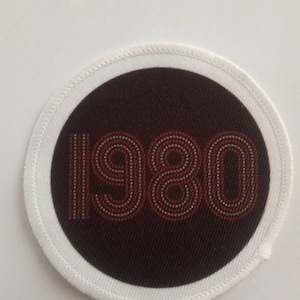 May include: A white circular patch with a black circle in the center. The black circle has the number 1980 printed in red.