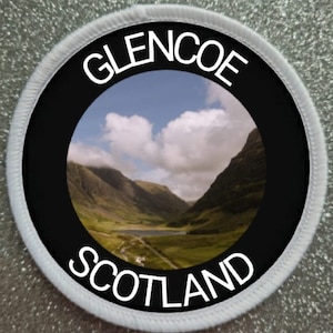 May include: A white round patch with black text that reads "Glencoe Scotland" and a photo of a valley with mountains and clouds in the centre.