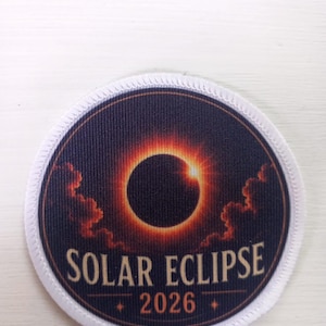 Puede incluir: Un parche bordado circular que muestra un eclipse solar oscuro rodeado por una corona naranja brillante y nubes. El texto "SOLAR ECLIPSE" está impreso en una fuente serif sobre el año "2026" con pequeños detalles de estrellas.