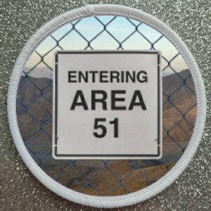 Puede incluir: Un parche circular blanco con un borde blanco. El parche tiene una foto de una valla de cadena con un letrero que dice "ENTERING AREA 51".