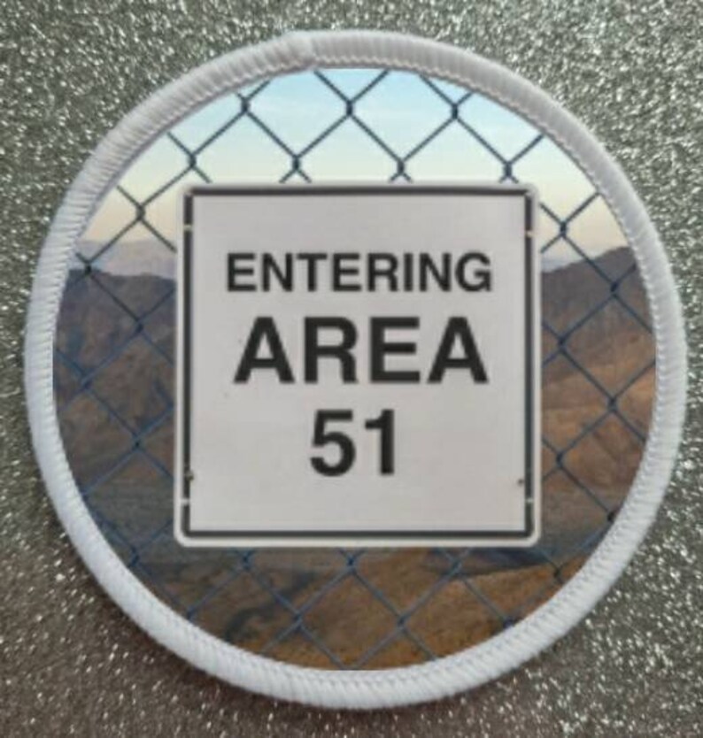 Pu&ograve; includere: Un cerotto circolare bianco con un bordo bianco. Il cerotto ha una foto di una recinzione a maglie con un cartello che recita "ENTERING AREA 51".