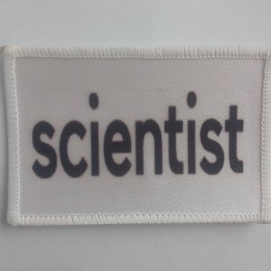 May include: White fabric patch with black stitching around the edges. The word "scientist" is printed in black letters on the patch.