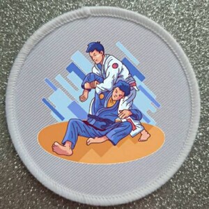 Puede incluir: Un parche redondo blanco con un patrón geométrico azul y blanco en el fondo. Dos figuras en uniformes de artes marciales azul y blanco se muestran en una posición de grappling. La figura de arriba lleva un uniforme blanco y la figura de abajo lleva un uniforme azul.