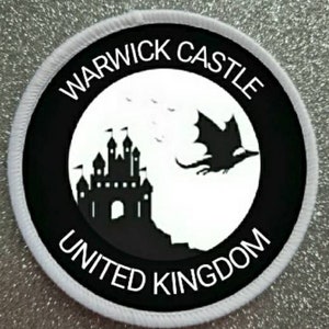 Puede incluir: Un parche circular blanco y negro con el texto "Warwick Castle United Kingdom" alrededor del borde. El centro del parche presenta una silueta negra de un castillo y un dragón.
