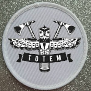 Peut inclure: Un patch rond blanc avec une image noire et blanche d'un totem avec deux haches de chaque côté. Le totem a une tête d'oiseau et des ailes. Le texte "TOTEM" est écrit en noir sous le totem.