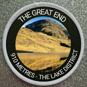 May include: A white round patch with black text and a photo of a mountain and lake. The text reads "The Great End 970 Metres - The Lake District".