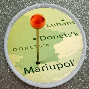 May include: A white circular patch with a map of eastern Ukraine. The map shows the cities of Luhansk, Donetsk, and Mariupol. A red push pin is placed on the map, marking the location of Mariupol.