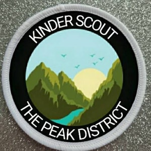 Op de afbeelding: Ronde witte patch met zwarte tekst die "Kinder Scout The Peak District" luidt. De patch toont een groene bergketen met een blauwe rivier en een gele zon op de achtergrond.