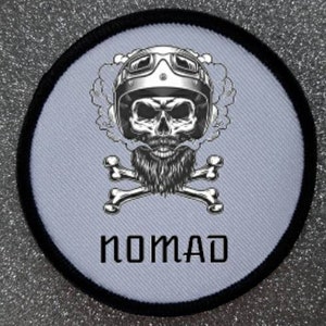 Könnte beinhalten: Ein weißer runder Patch mit schwarzem Rand, der einen Totenkopf mit Motorradhelm, Bart und gekreuzten Knochen zeigt. Der Text "NOMAD" ist unter dem Totenkopf gedruckt.