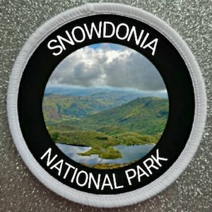 Może przedstawiać: Okrągła biała łatka z czarną obwódką i białym tekstem, który brzmi "Snowdonia National Park". Środek łatki przedstawia zdjęcie pasma górskiego z jeziorem na pierwszym planie.