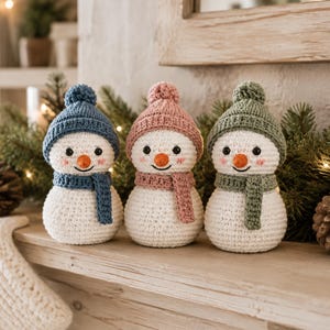 May include: Three crocheted snowman figurines, each featuring a white body, a small orange nose, black eyes, and pink cheeks. Each snowman wears a matching colored beanie with a pom-pom and a scarf in blue, dusty pink, or sage green.