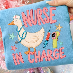 May include: Light blue t-shirt with a cartoon duck wearing a nurse's hat and stethoscope. The shirt features the words "NURSE IN CHARGE" in pink, along with illustrations of a syringe, thermometer, and stars.