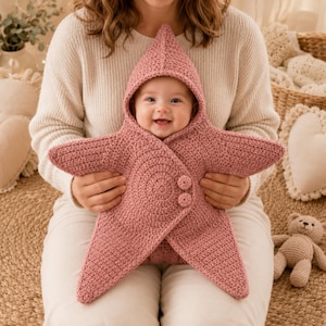 May include: A dusty rose crochet baby swaddle blanket shaped like a five-pointed star with a pointed hood. The garment features a textured knit pattern and two matching circular buttons on the front flap to secure the wrap around the infant.