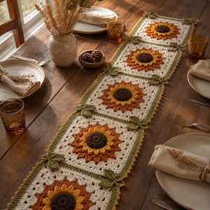 May include: A crochet table runner featuring a series of square panels, each displaying a three-dimensional sunflower with a dark brown center, orange and yellow petals, and cream-colored borders, accented with small green crocheted leaves.
