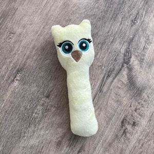 以下が含まれることがあります： 淡い黄色をした、柔らかいフクロウの形をしたおもちゃ。フクロウは、ターコイズ色の虹彩、黒いまつげ、茶色のくちばしを持つ大きな刺繍の目を持っています。おもちゃは長く円筒形の体でデザインされています。