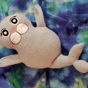 May include: A plush gray seal toy with embroidered facial features, including large eyes, a pink nose, and whiskers. The seal is lying on a blue and green tie-dye patterned fabric. The toy has flippers and a tail.