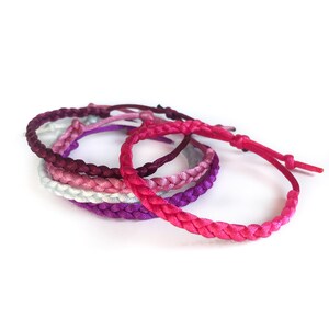 May include: A set of five braided bracelets in shades of pink, purple, and white. The bracelets are made of a soft, silky material and are tied together in a knot.