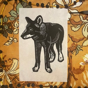 May include: Two black and white linocut prints of a wolf on a patterned fabric background. The prints are on separate pieces of paper and are framed by a brown fabric border.