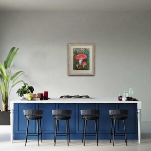 May include: A framed painting of red and white mushrooms in a forest setting. The painting is in a light wood frame and is hanging on a white wall.
