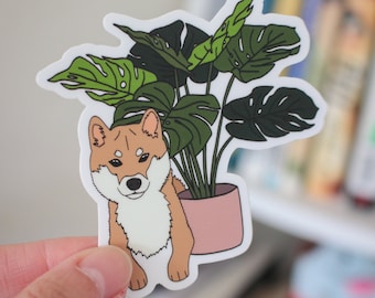 May include: A sticker featuring a cartoon Shiba Inu dog peeking out from behind a potted Monstera plant with green leaves. The dog is brown and white with a pink nose and the pot is pink.