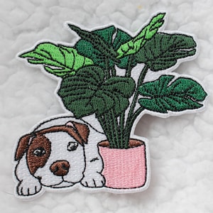 May include: Embroidered patch of a white dog with brown spots peeking out from behind a potted green plant with pink pot.
