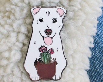 May include: White enamel pin of a dog holding a potted cactus with pink flowers. The pin is on a denim jacket with a white sherpa collar.
