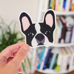 May include: A black and white illustrated sticker of a French bulldog's face. The sticker has a white background and pink ears.