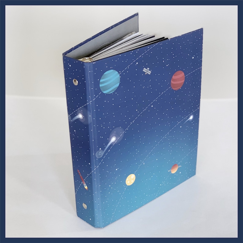 May include: A three-ring binder with a space-themed design. The cover features a dark blue background with illustrations of planets, stars, a rocket, and a satellite. The binder is open, revealing white paper inside.