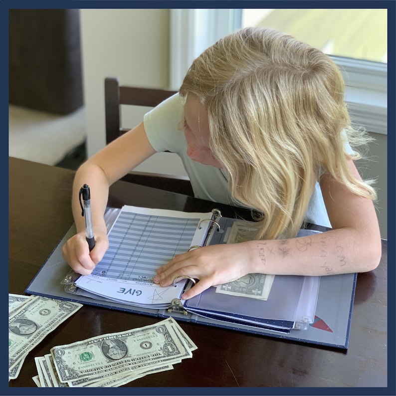 May include: A young person writing in a blue binder labeled "GIVE" with a pen. Several one-dollar bills are scattered on the table. The binder contains clear pockets with money inside. The person has blonde hair and is wearing a light green shirt.