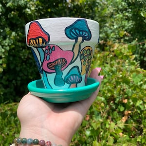 May include: A white ceramic planter with a green saucer, hand-painted with colorful mushrooms. The planter has a whimsical design with various mushroom shapes and colors.