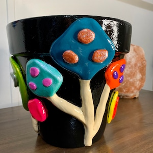 May include: Black ceramic planter with colorful, whimsical mushroom-shaped decorations. The mushrooms are painted in shades of blue, green, pink, orange, and yellow.