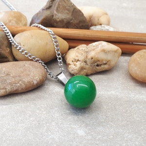 May include: A silver chain necklace with a bright green, round pendant. The necklace is displayed on a gray surface with small stones and wooden sticks.