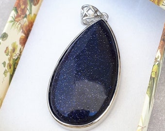 May include: A dark blue glitter stone pendant in a teardrop shape, set in a silver-coloured metal frame. The pendant is presented in an open gift box with a floral pattern and a white interior.