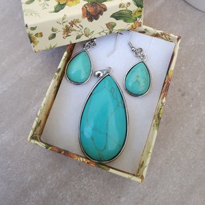 Puede incluir: Un conjunto de joyas turquesas, que incluye un colgante y pendientes, presentado en una caja de regalo con estampado floral. Las piedras en forma de l&aacute;grima est&aacute;n engastadas en metal plateado. La caja est&aacute; abierta, revelando las joyas.