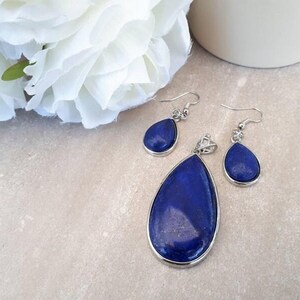 Pu&ograve; includere: Un set di gioielli con un ciondolo e orecchini abbinati. Il set ha una pietra blu scuro a forma di goccia incastonata in metallo color argento. Il ciondolo &egrave; pi&ugrave; grande degli orecchini. Un fiore bianco e una tazza di ceramica bianca sullo sfondo.