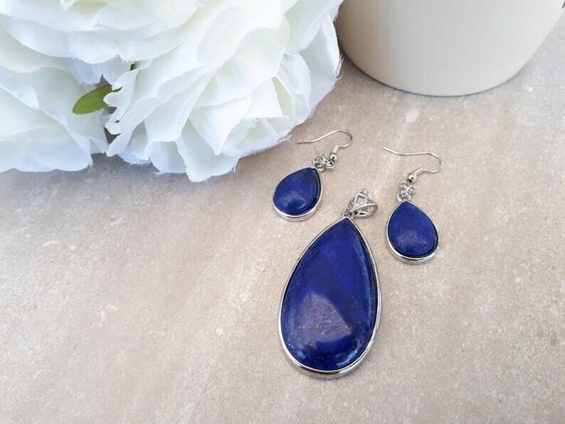 Pu&ograve; includere: Un set di gioielli con un ciondolo e orecchini abbinati. Il set ha una pietra blu scuro a forma di goccia incastonata in metallo color argento. Il ciondolo &egrave; pi&ugrave; grande degli orecchini. Un fiore bianco e una tazza di ceramica bianca sullo sfondo.